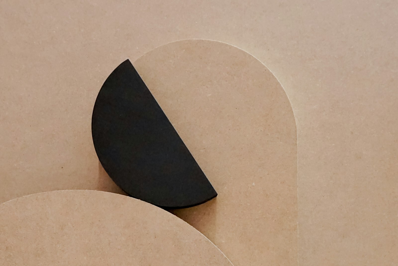 A matte black paper half circle resting against pale tan board cut into arcs, flat geometry with nothing else in the frame