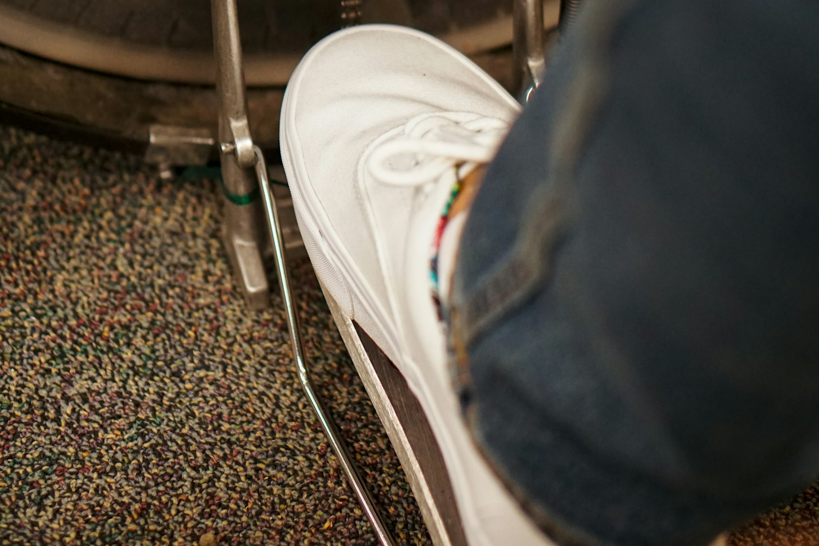 A foot in a white sneaker resting on the footboard of a kick drum pedal, the bass drum hoop visible behind it on a patterned venue carpet
