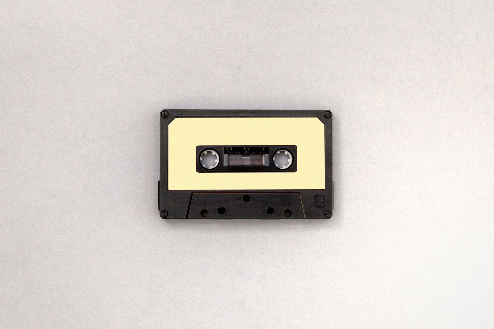 A single black and cream audio cassette lying flat on a plain pale grey surface, the tape format lofi takes its texture from