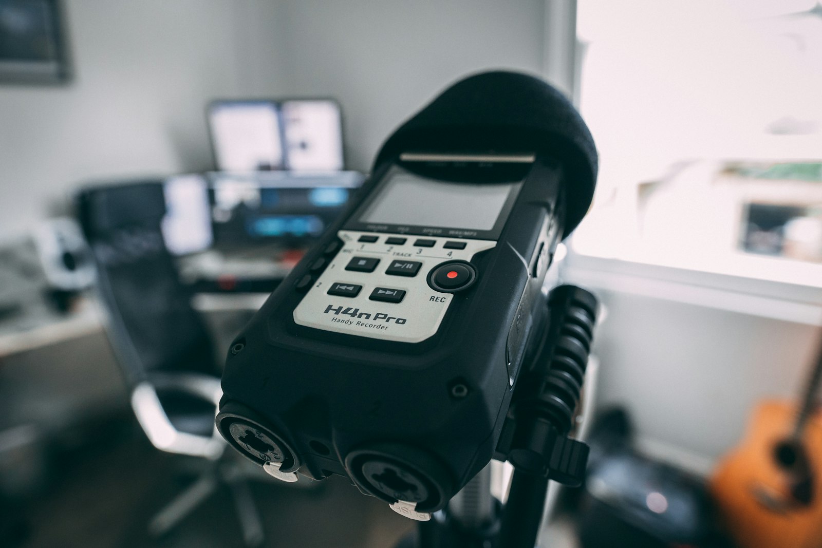 A Zoom H4n Pro handy recorder mounted on a stand in a home studio with a laptop and an acoustic guitar blurred behind it, the kind of device a recording arrives from
