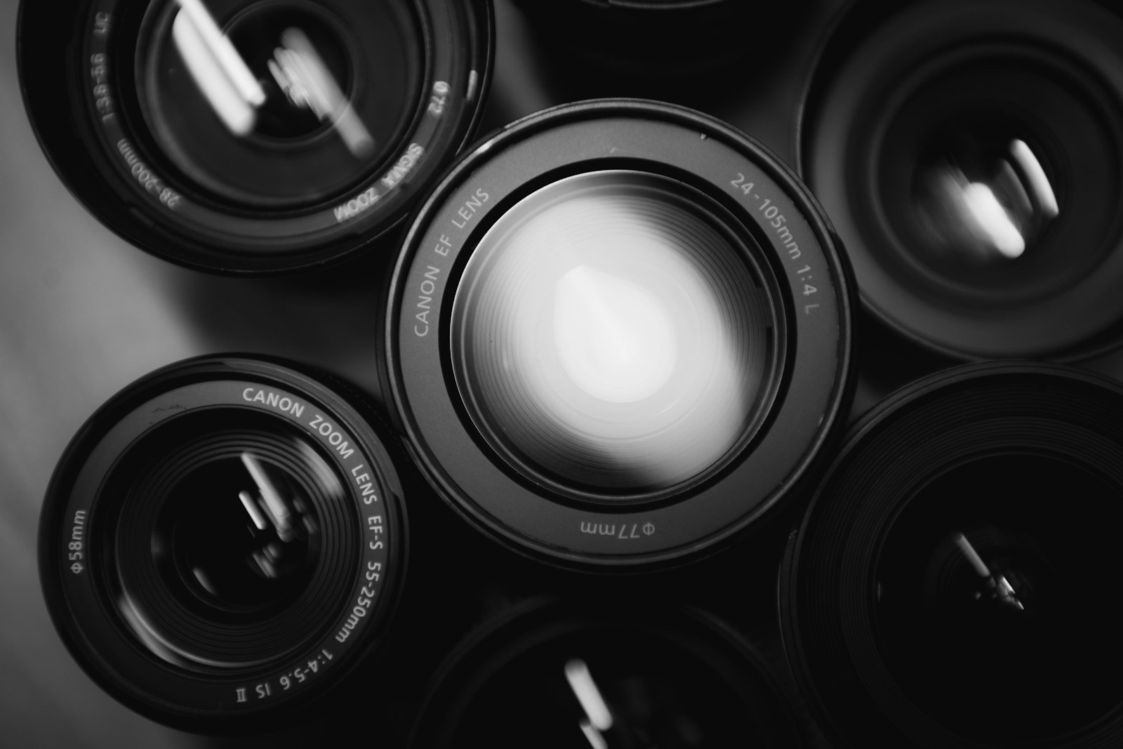 Six camera lenses photographed from above in black and white, packed together with one of them catching a bright highlight while the rest stay dark