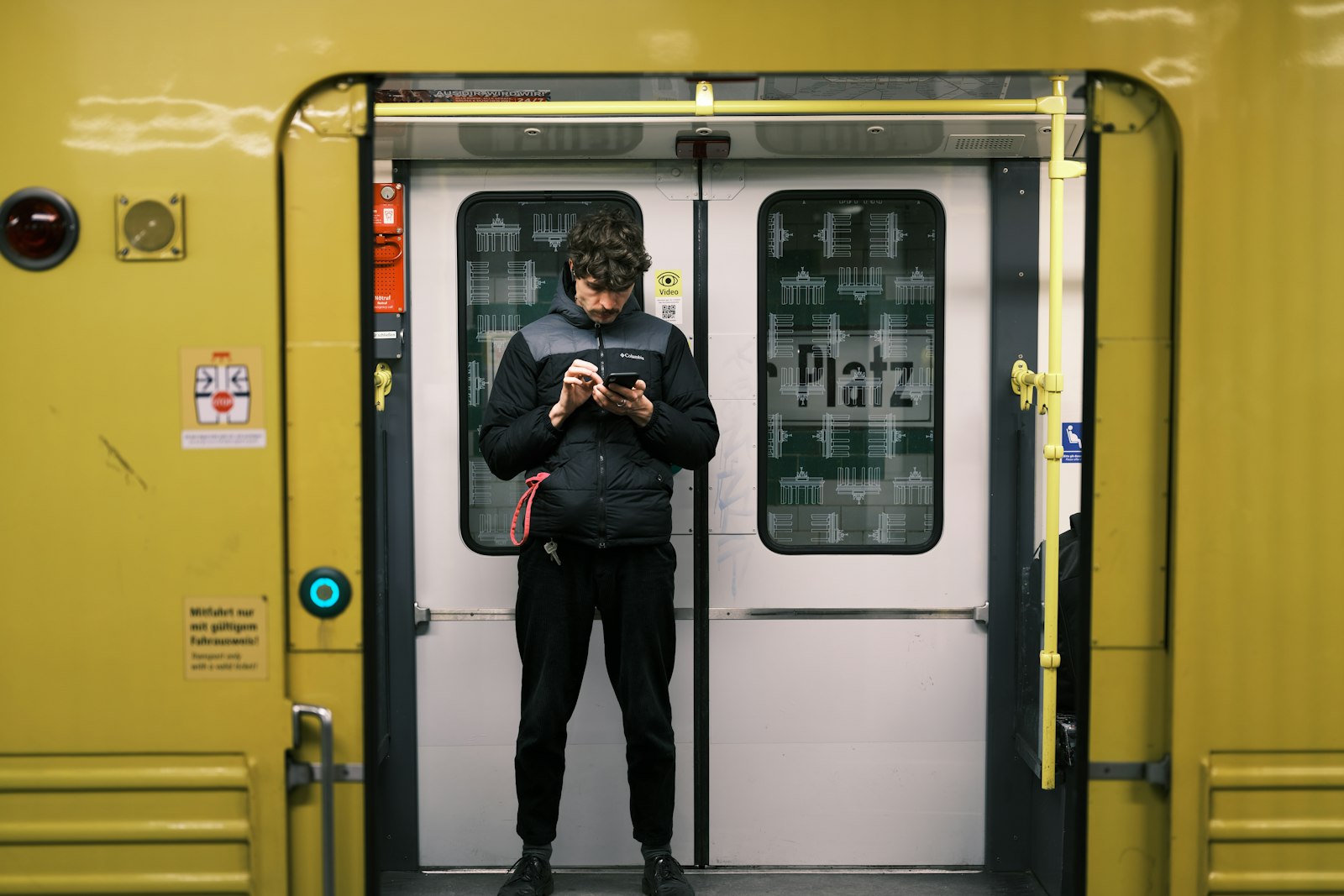 A man standing in the doorway of a yellow subway train looking down at his phone with no headphones in