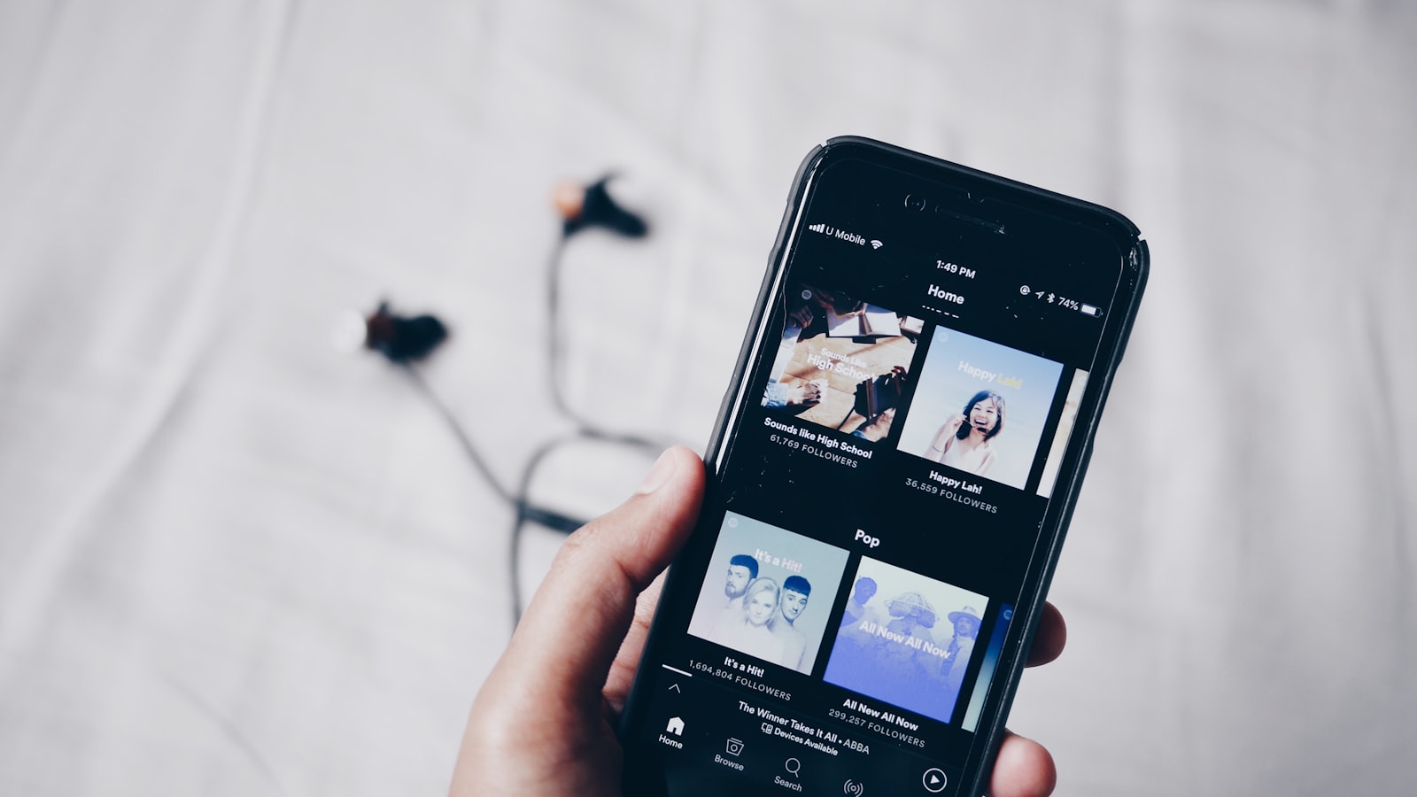 A hand holding a phone with a music app open on the screen and a pair of earbuds lying on the sheet behind it