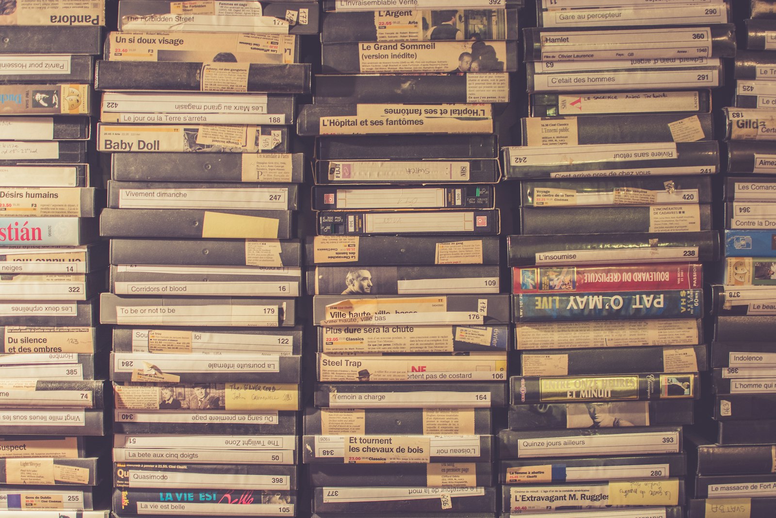 A wall of hand labelled VHS tapes stacked spine out, their paper labels typed and handwritten, the tape format the artifacts layer is imitating