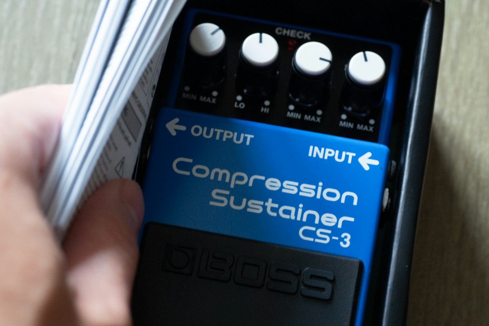 A hand holding a Boss CS-3 Compression Sustainer guitar pedal, its four white knobs sitting above MIN and MAX scales printed on the chassis